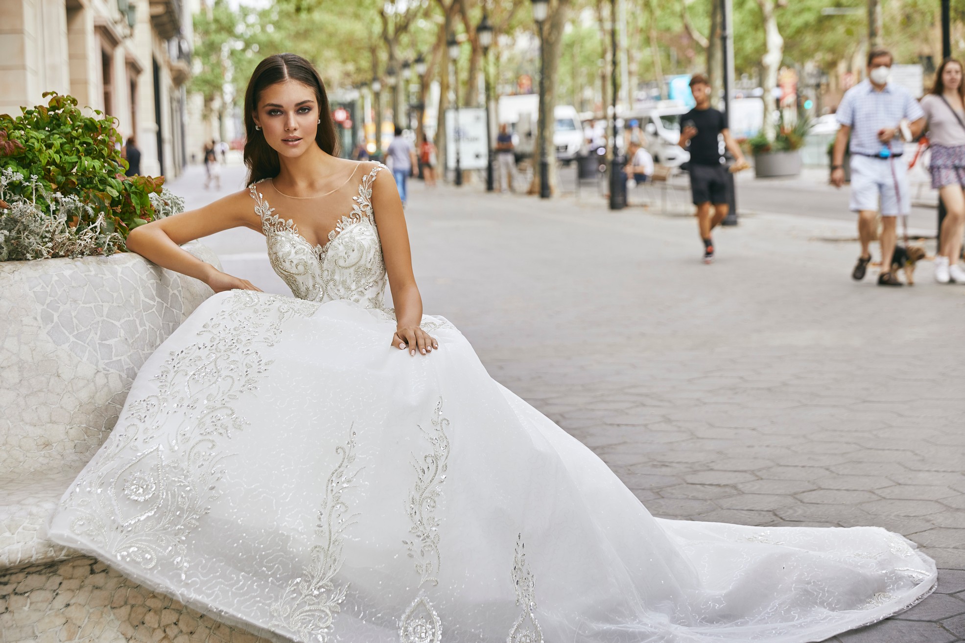 Brunette model sat in a tree-lined sunny street in Ronald Joyce 69720, an ivory A-line wedding dress style with an illusion sweetheart neckline, silver beaded bodice and illusion off-the-shoulder straps